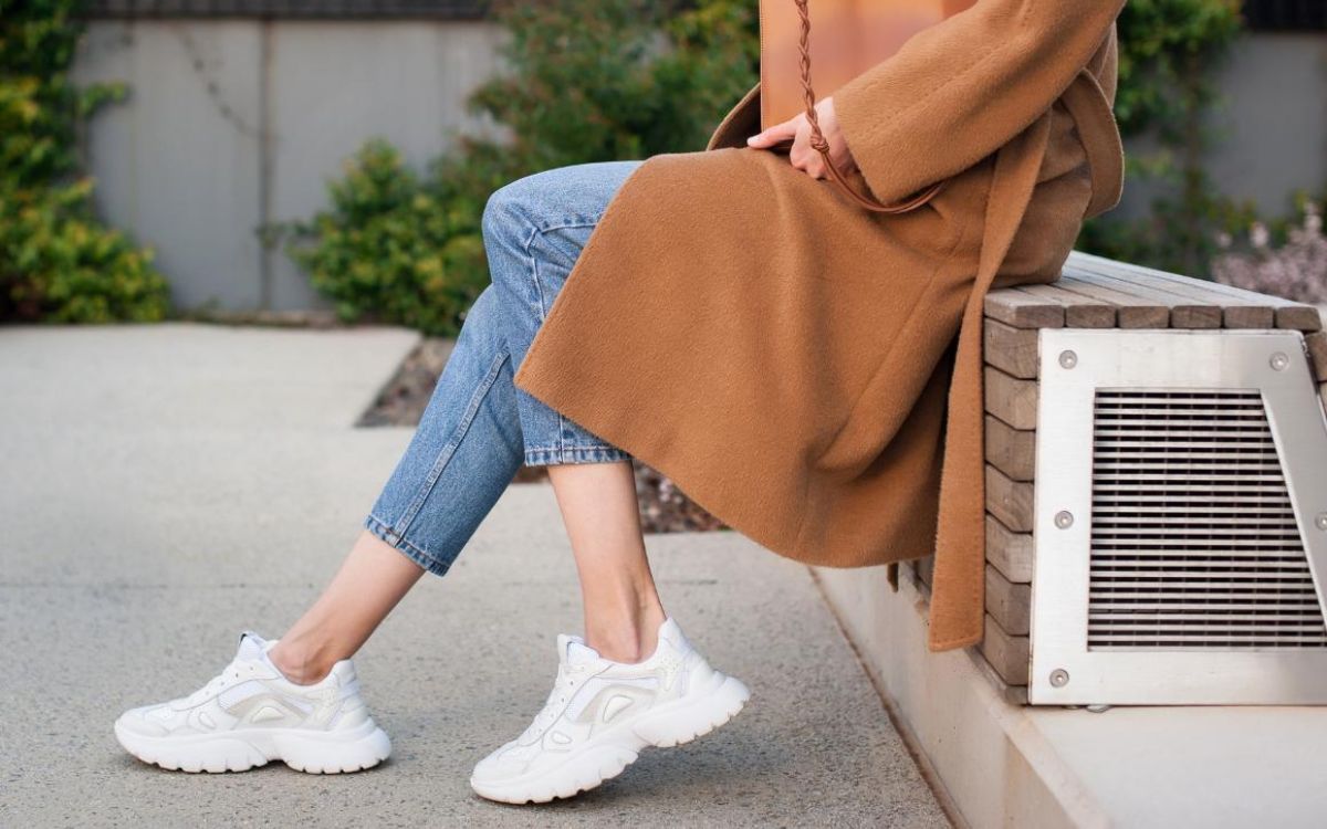 Mulher sentada, usando casaco longo marrom, calça jeans e tênis branco