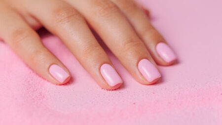 Mão feminina com unhas curtas e esmaltadas em tom rosa claro sobre fundo rosa, representando o resultado dos cuidados para fortalecer as unhas e mantê-las saudáveis.
