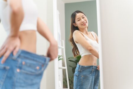 Calças femininas: guia completo dos principais modelos e como usar Mulher sorrindo em frente ao espelho vestindo calças femininas jeans largas, ajustando o cós e observando o caimento da peça.