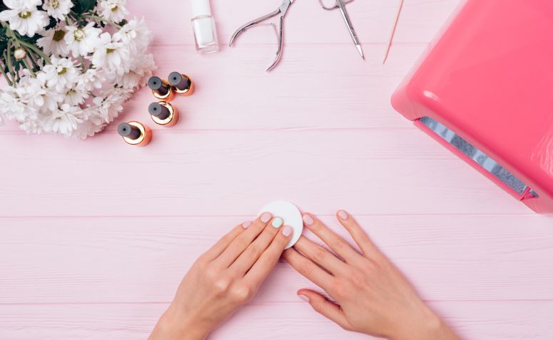Mãos femininas removendo esmalte em gel com algodão sobre as unhas, acompanhadas de itens de manicure e frascos de esmalte sobre uma mesa rosa — processo de uso de removedor de esmalte em gel de forma delicada e segura.
